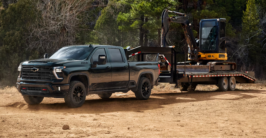 Black Chevrolet Silverado HD Trail Boss towing an excavator on a flatbed trailer across a dirt lot