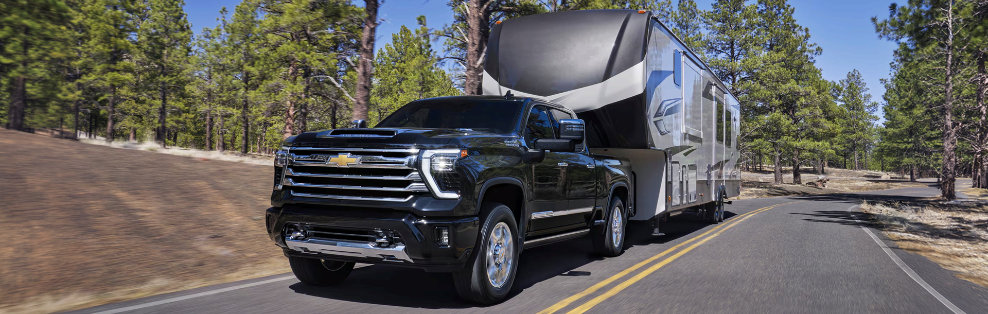 Black Chevrolet Silverado HD towing a fifth-wheel camper along a pine-lined road