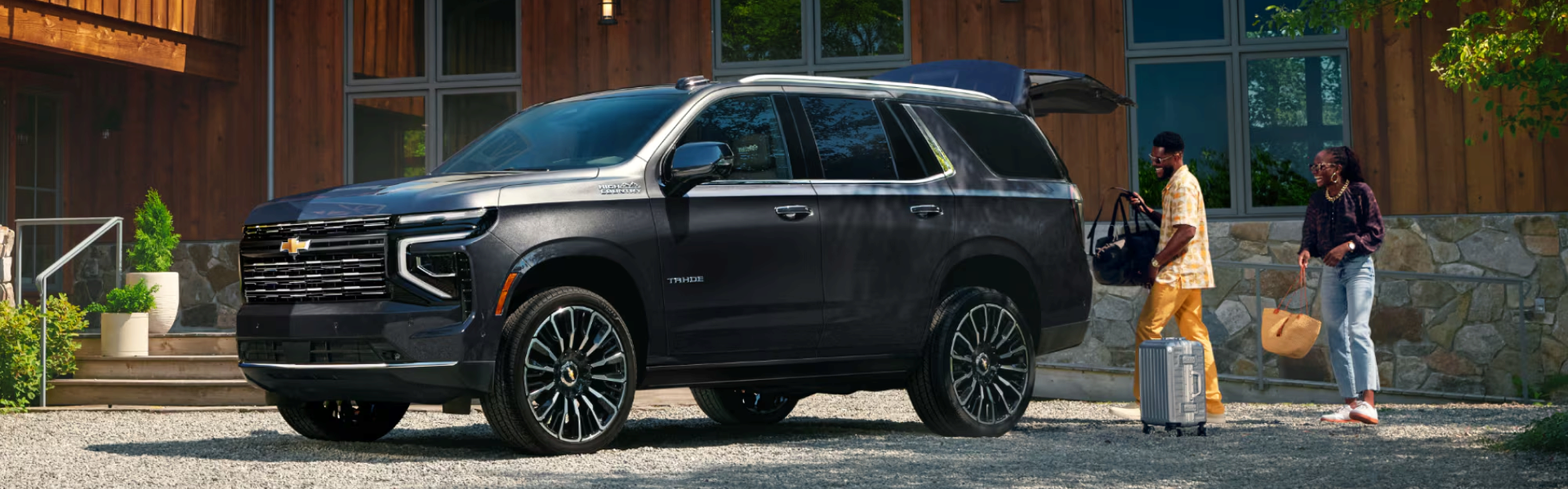 Black 2027 Chevrolet Tahoe with its liftgate open as a couple loads luggage outside a lodge