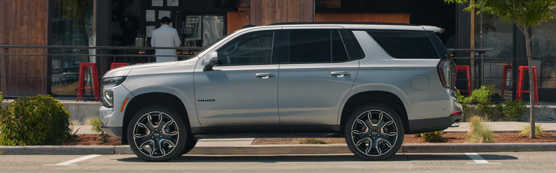 Silver 2027 Chevrolet Tahoe parked at the curb outside a café, shown in profile