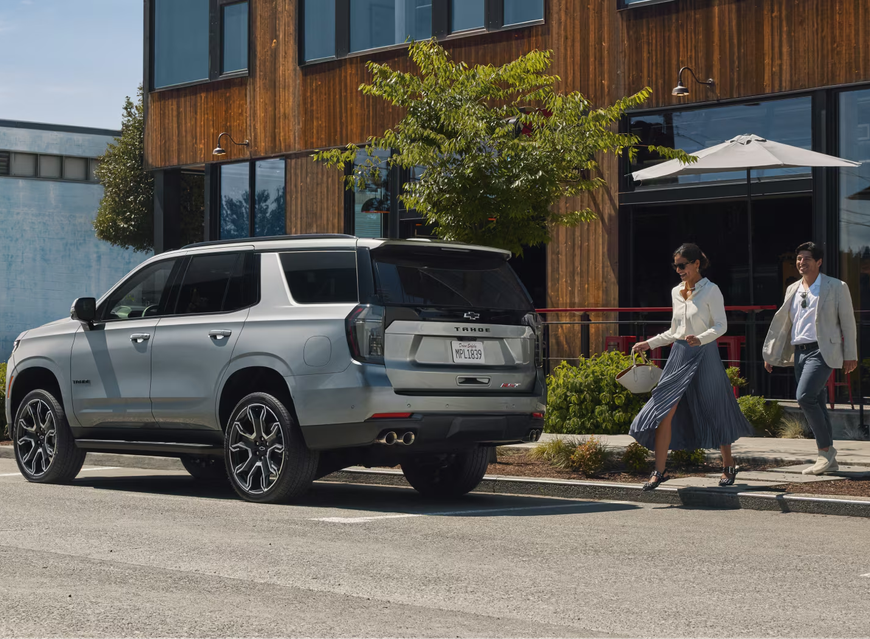 Silver Chevrolet Tahoe parked on a street as two people walk past a modern wood-clad building