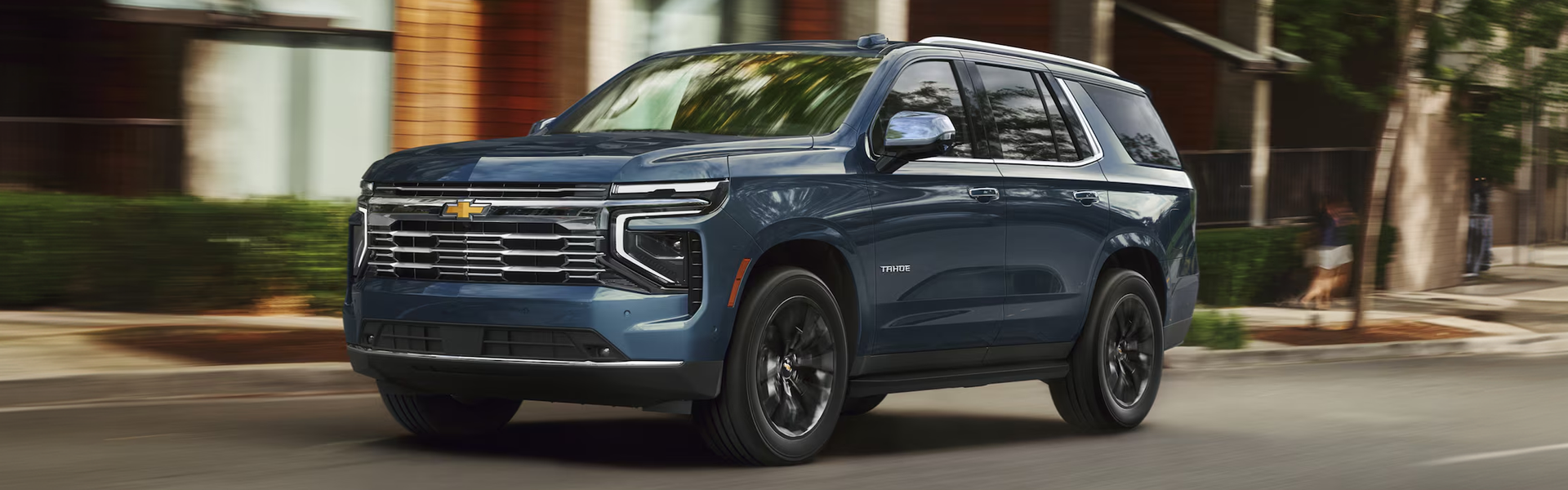 Blue 2027 Chevrolet Tahoe driving down a tree-lined city street