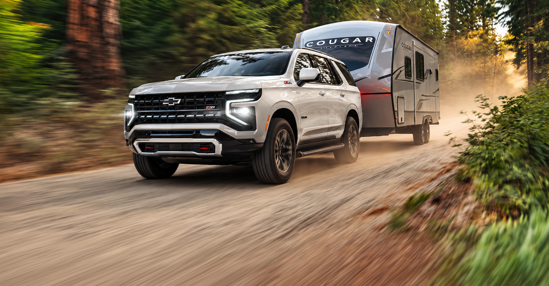 White 2027 Chevrolet Tahoe towing a travel trailer down a dusty forest road