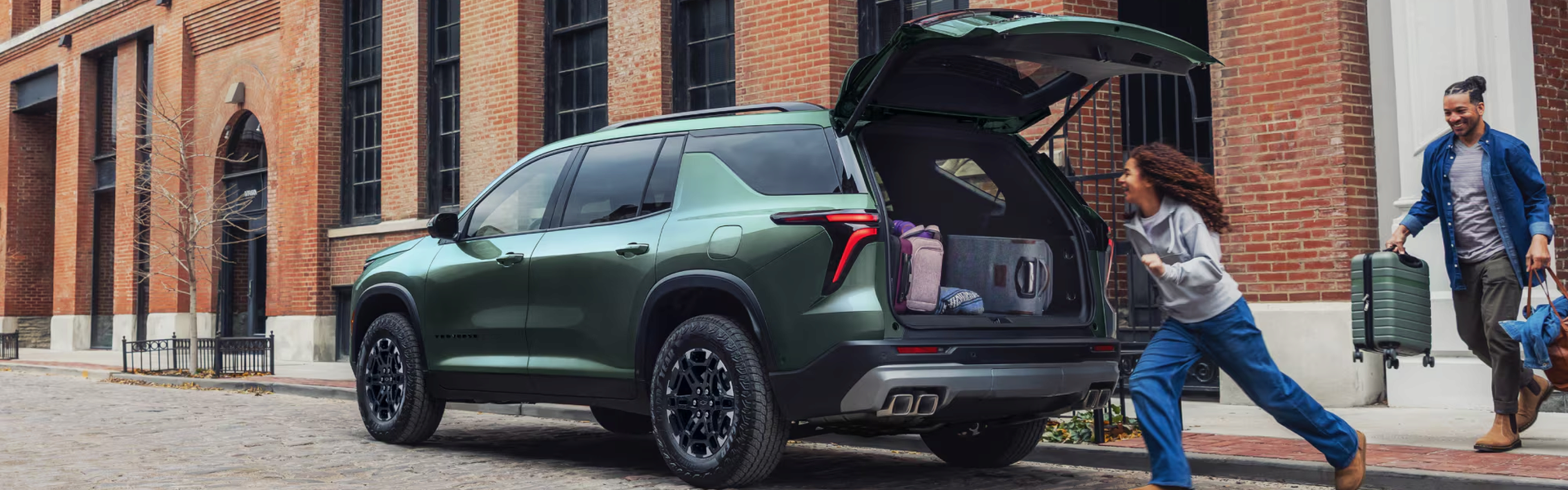 Green Chevrolet Traverse with its liftgate open as a family loads luggage on a city street