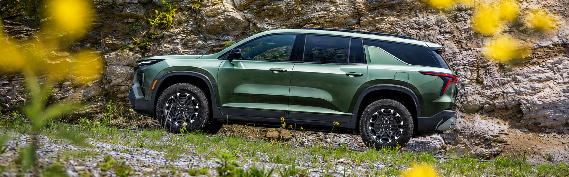 Green 2027 Chevrolet Traverse driving past a rocky hillside with yellow wildflowers