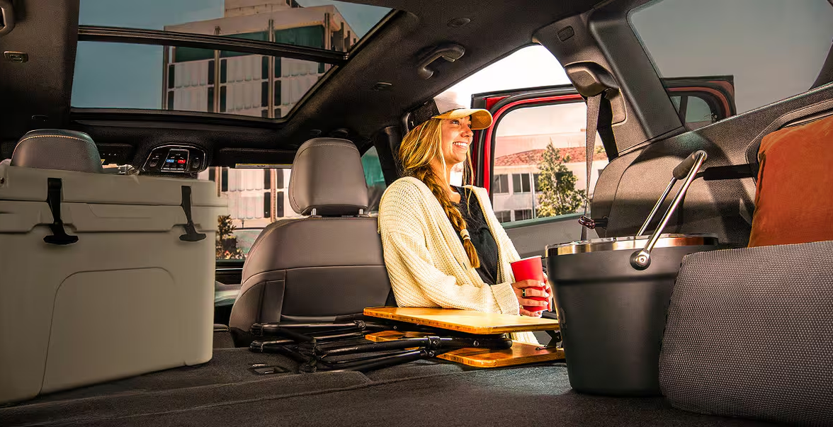 Woman relaxing in the open cargo area of a 2027 Jeep Cherokee packed with a cooler and camping gear