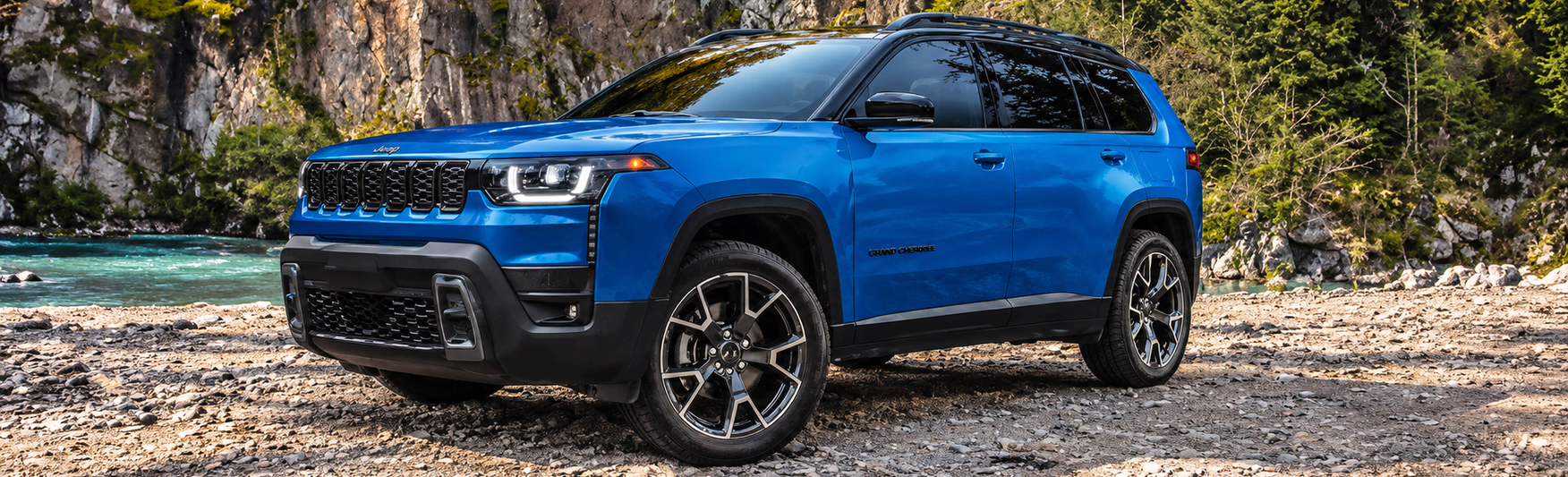 Blue 2027 Jeep Cherokee parked on a rocky riverbank below cliffs
