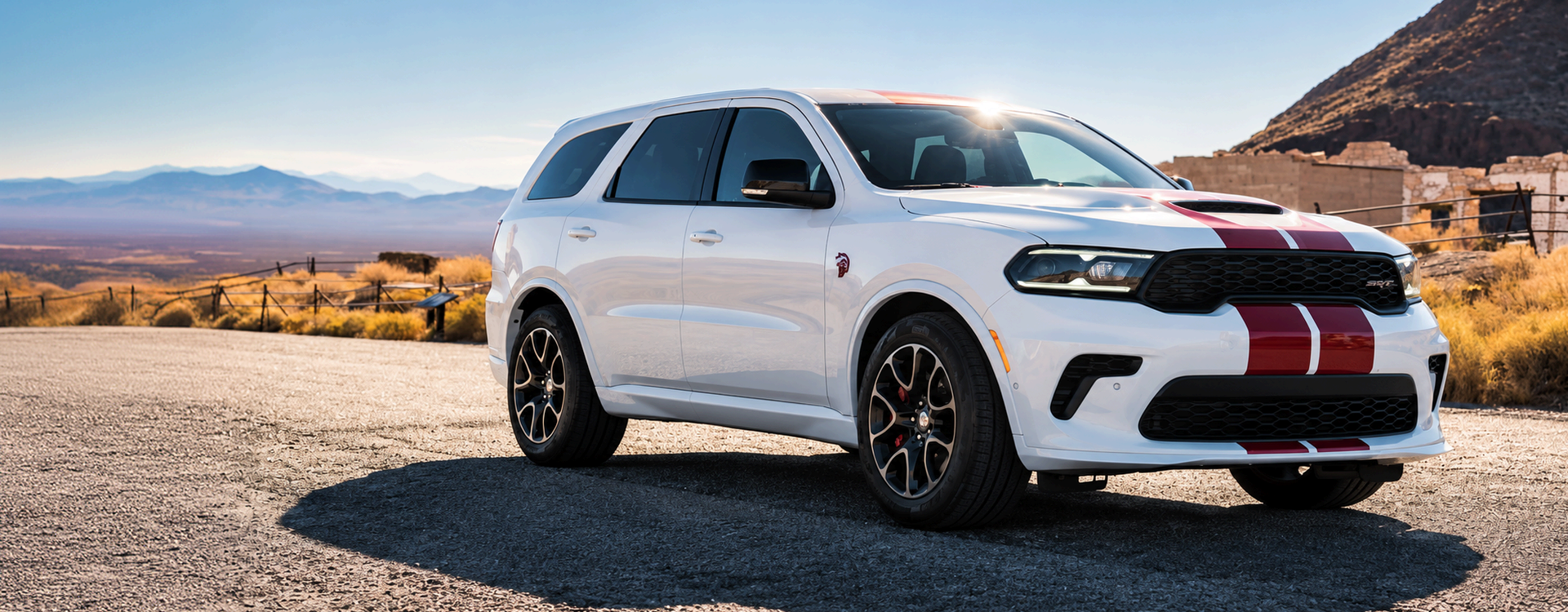 White 2027 Dodge Durango SRT with red racing stripes parked on gravel near desert ruins and a rocky hillside