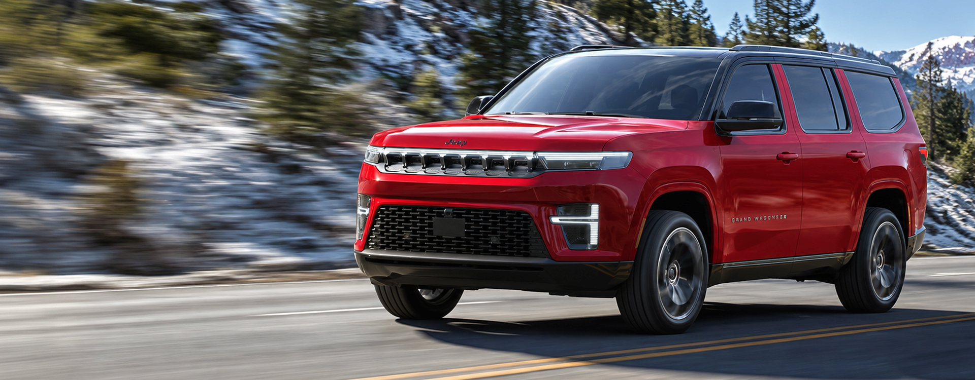 Red 2027 Jeep Grand Wagoneer driving on a mountain road past snowy pines